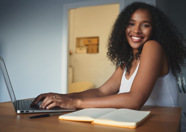 People, lifestyle, modern technology, job and communication concept. Attractive young dark skinned female freelancer with curly hair having pleased look, enjoying distant work, using generic laptop